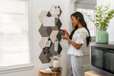 A Felt Right wall art design in shades of green, cream, and grey creating a 3d cube look that is pinnable. Next to a window and above a side table with a woman pinning card on it.