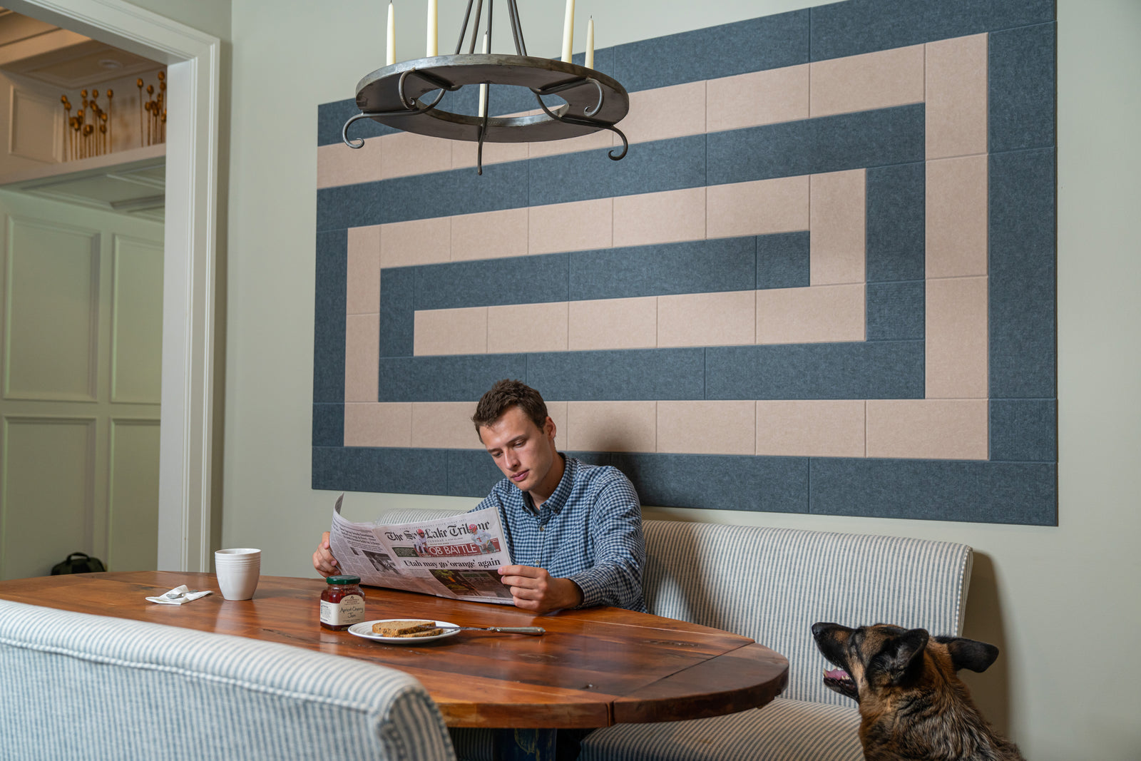 A Felt Right wall art design in shades of grey and tan felt tiles creating a spiral design on a green wall in a dinning room with a man reading the newspaper and his dog.