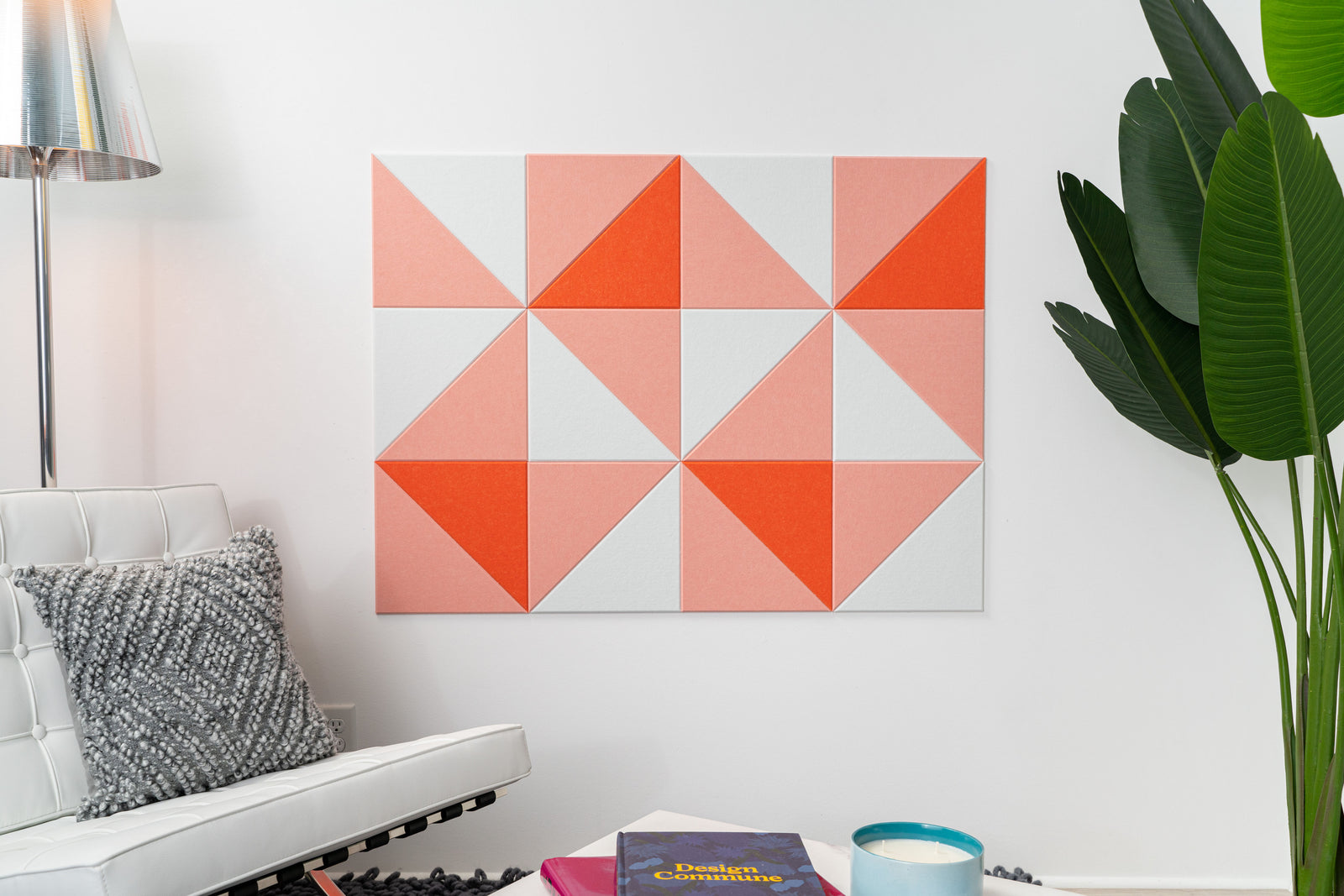 A Felt Right wall art design in shades of pink and white triangles creating geometric pattern on a white wall next to a chair and a house plant.