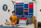 A Felt Right wall art design in shades of blue and red felt tiles as a headboard for a bed on a white wall.
