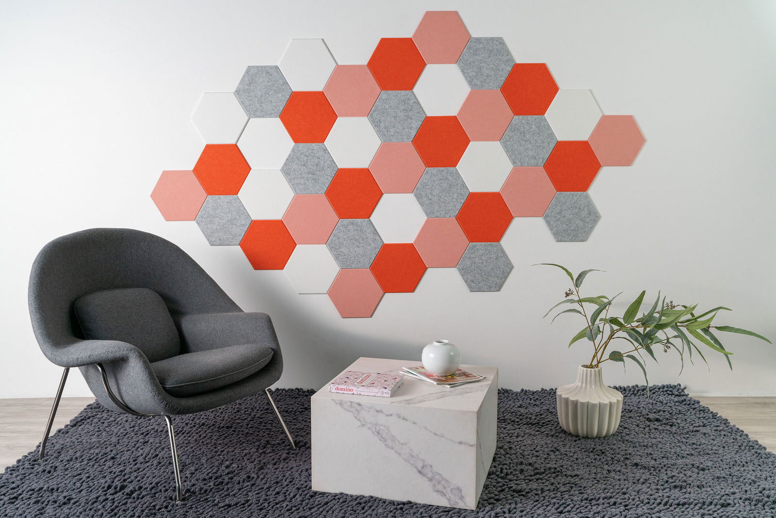 Felt Right wall art design using orange, pink, grey, and white hexagon felt tiles in a honeycomb pattern on a white wall by a chair, side tables, and a house plant