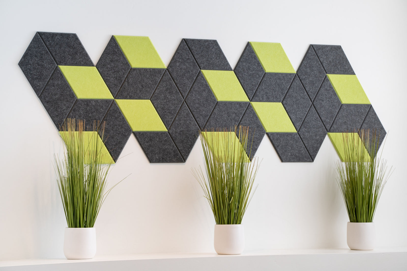A Felt Right Wall art design in grey and green creating a geometric shape along a white wall with three plants in front