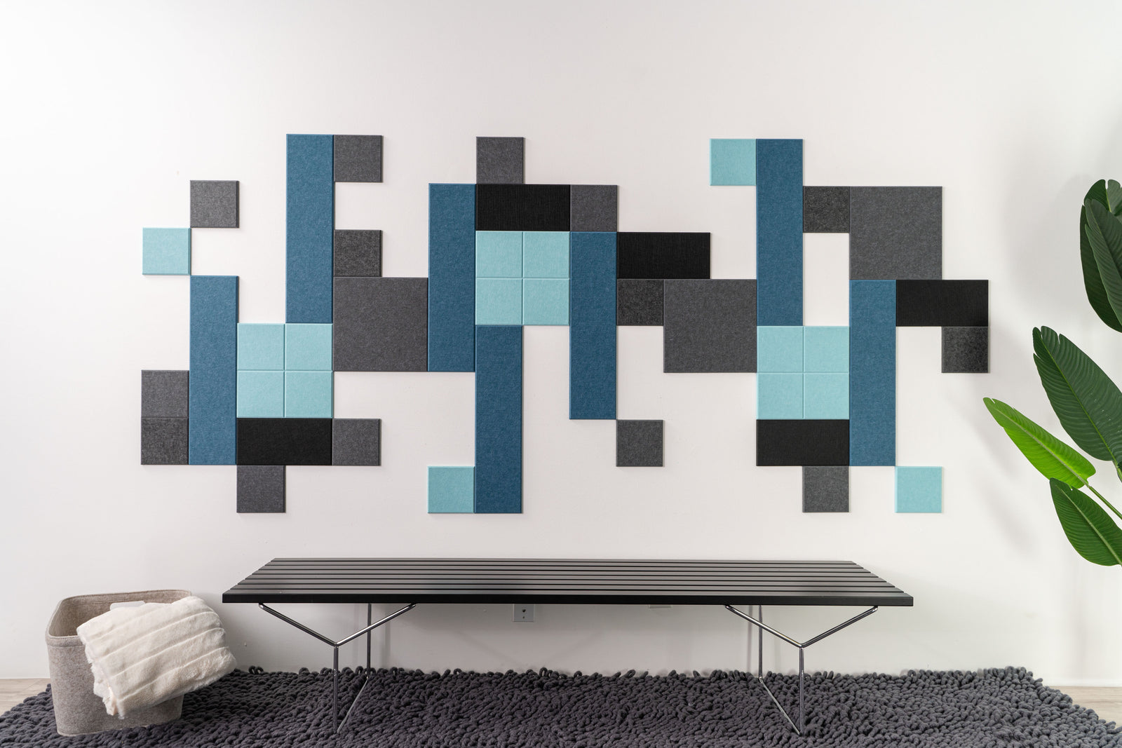A Felt Right wall art design using shades of blue and gray above a bench on a rug next to a house plant and a basket with a blanket