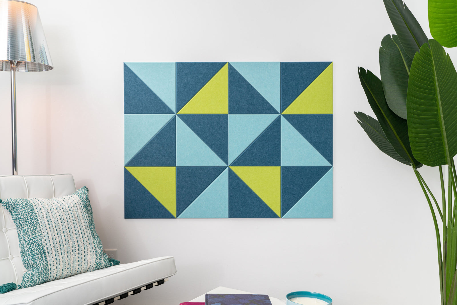 A Felt Right wall art design in shades of blue and green triangles creating a geometric wall art on a white wall next to a chair and a plant.