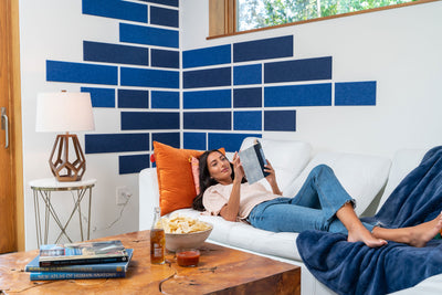 A Felt Right wall art design in shades of blue felt tiles spaced on a white wall with a woman on a couch reading.