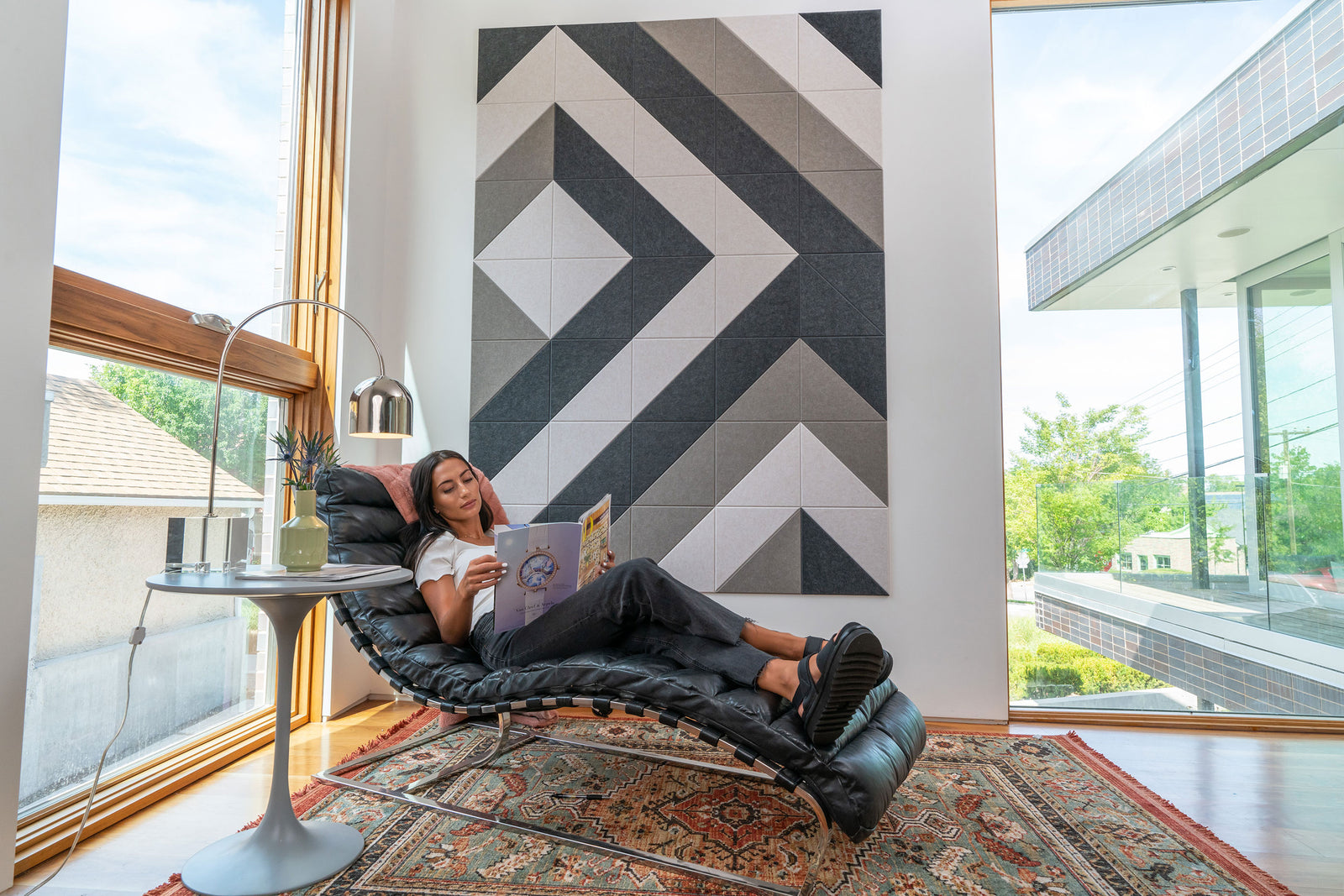A Felt Right wall art design in shades of cream and grey and brown on a white wall in between two big windows with a woman on a chair reading on a rug.