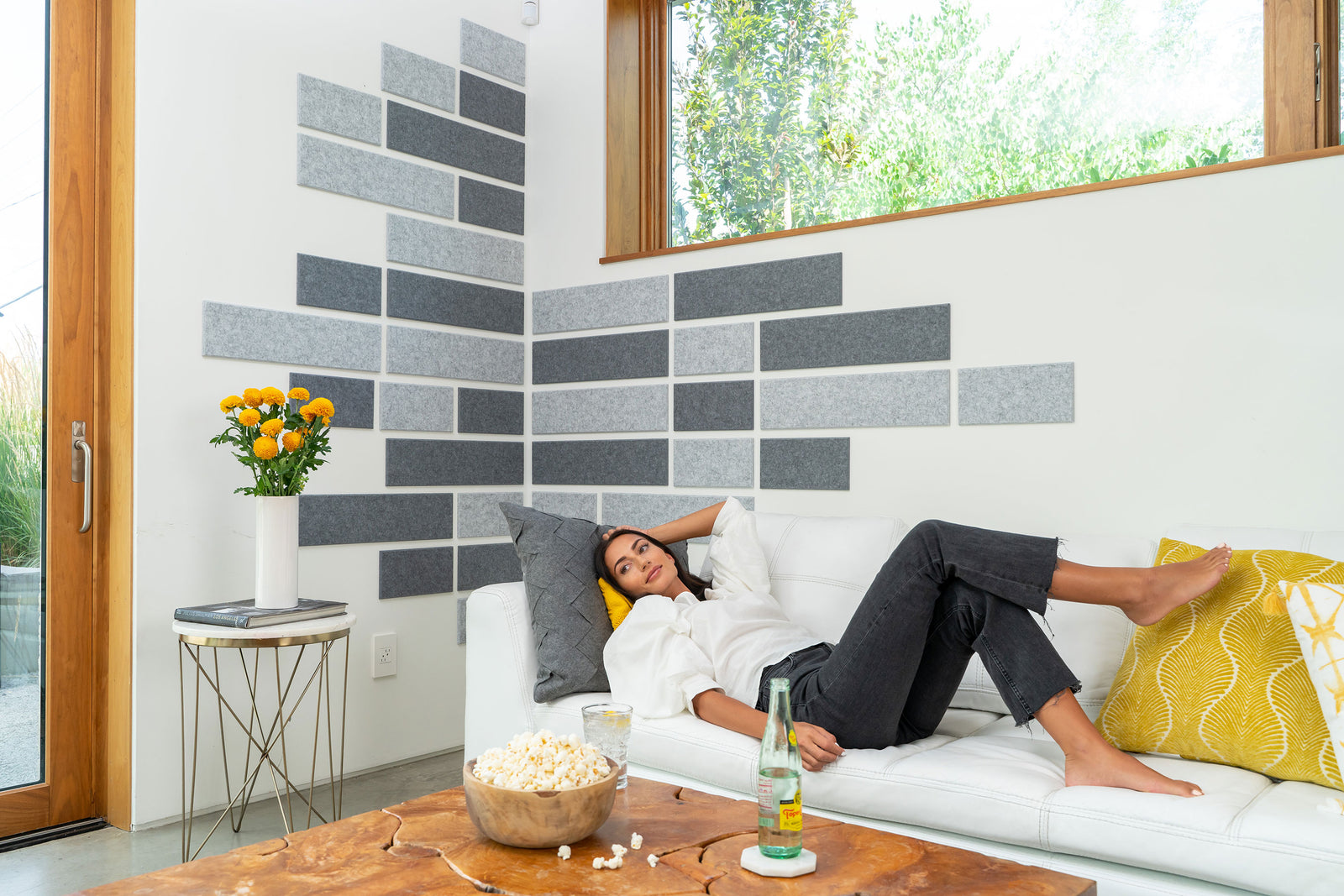 A Felt Right Wall art design in shades of grey felt tiles on a white wall corner with a woman sitting on a couch.