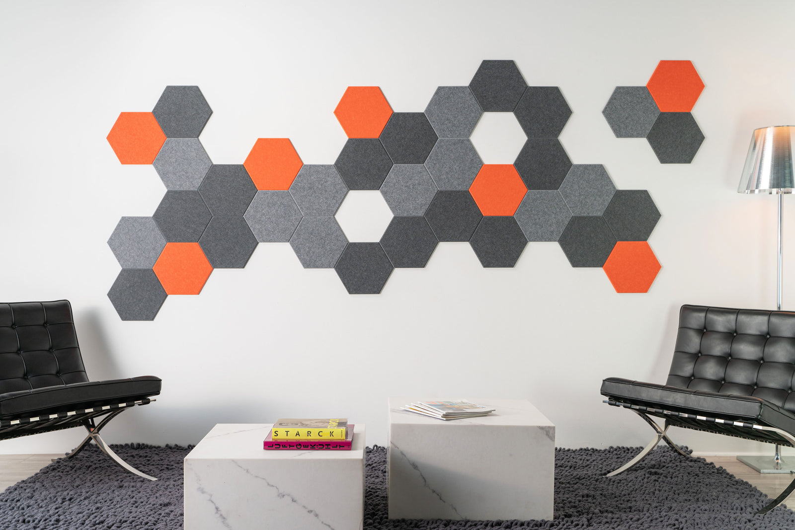 Felt Right wall design using hexigonal designs with pops of orange surrounded by shades of grey in a modern room with chairs and side tables.