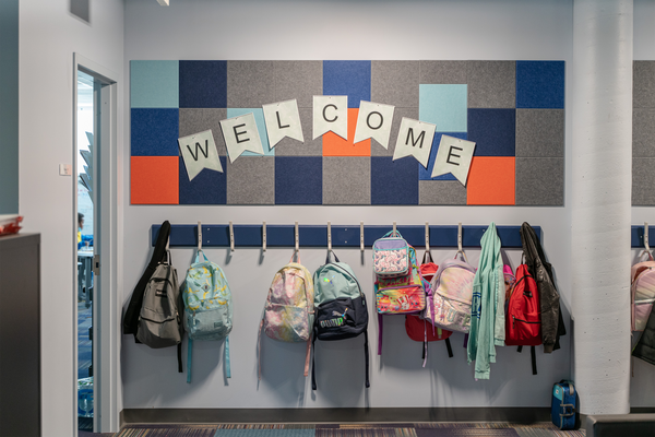 A Felt Right wall art design in shades of grey, blue, and orange square felt tiles with a pinnable welcome banner on a wall above a coat rack with backpacks in a classroom.