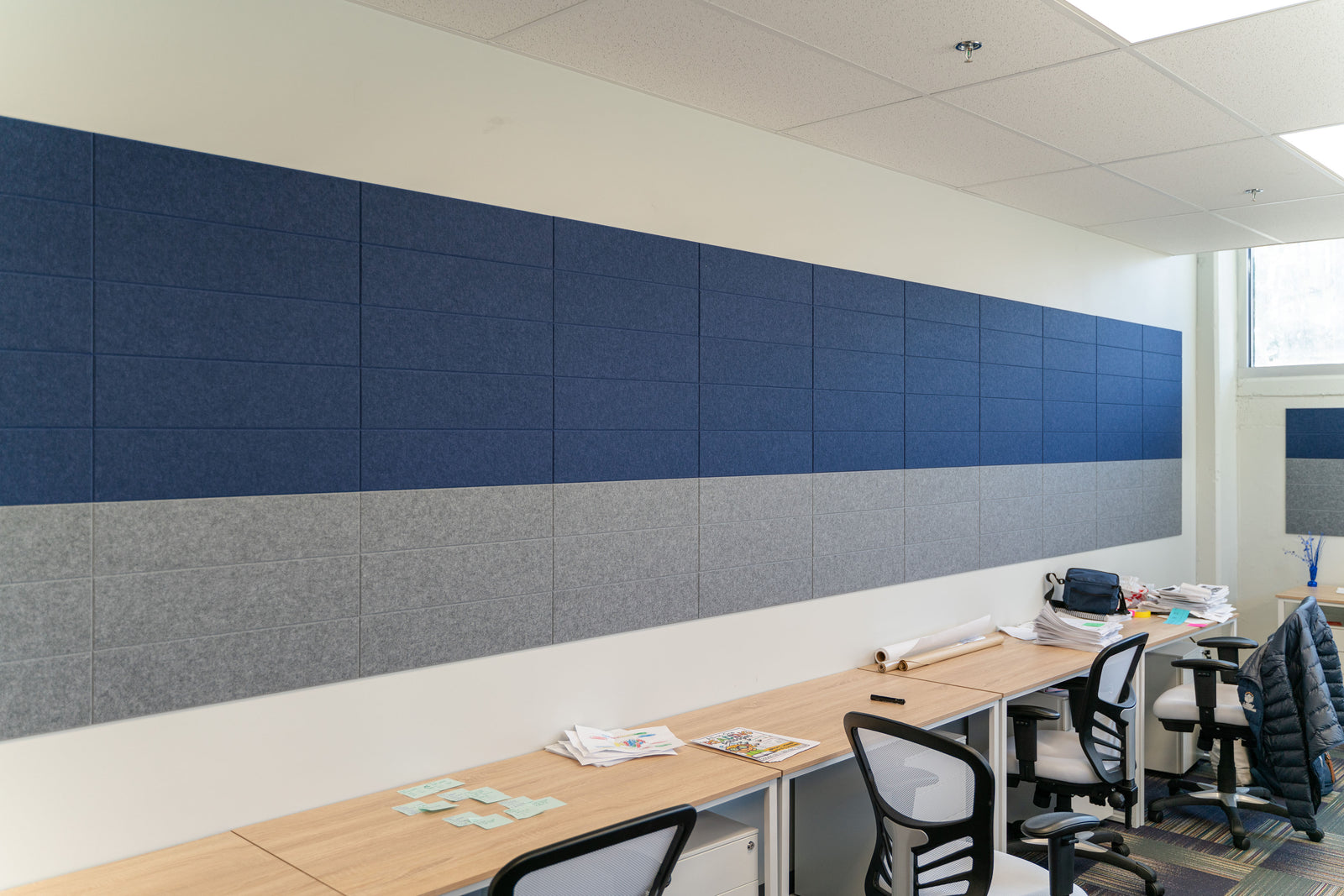 A Felt Right wall art design in a shiplap pattern using shades of blue and grey on a white wall in an office room with many desks.