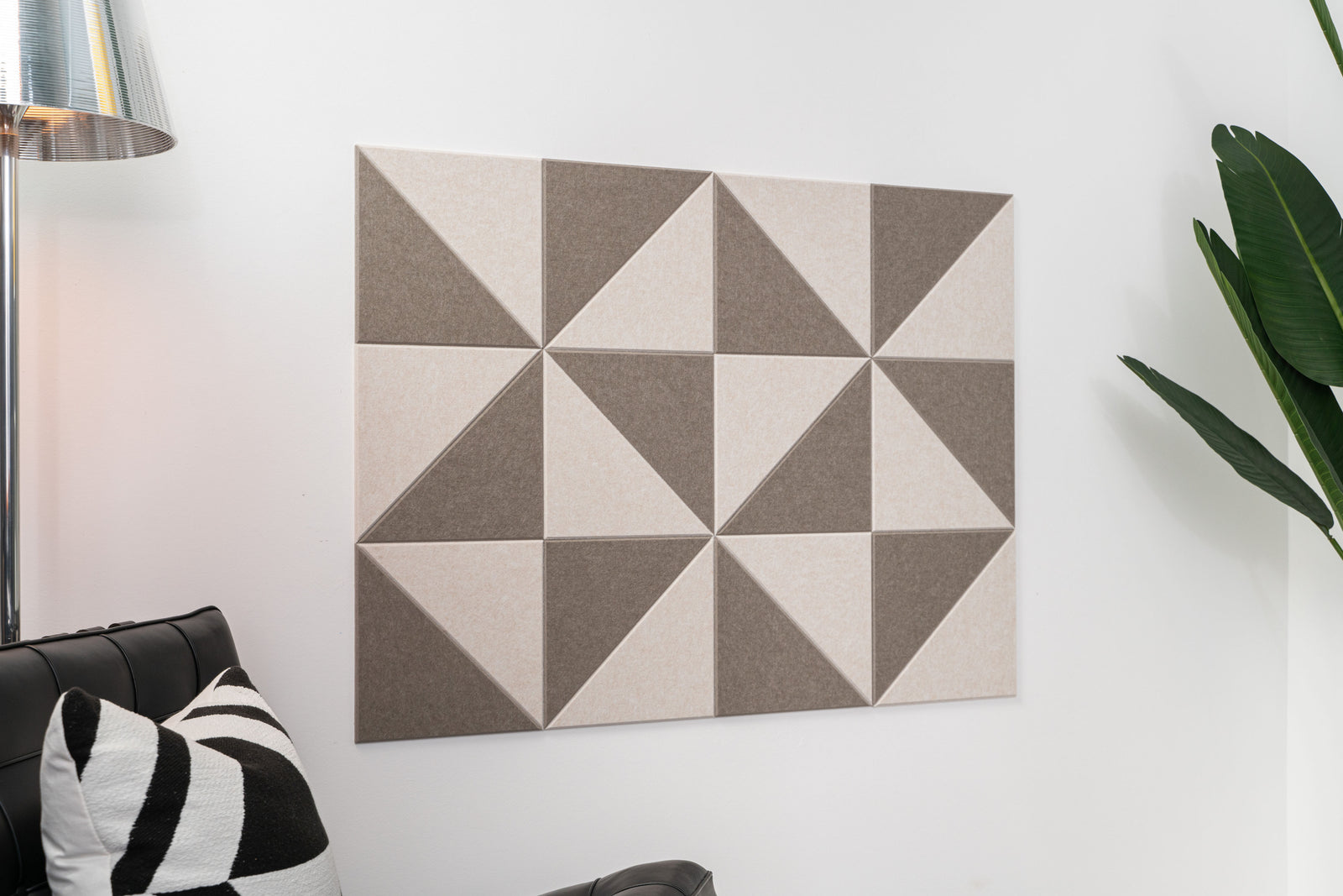 A Felt Right wall art design in shades of brown using triangles to create a pattern on a white wall next to a lamp and a chair with a decorative pillow.
