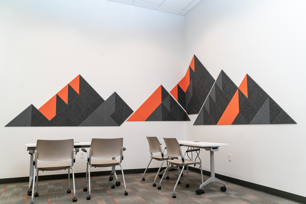 A Felt Right wall art design in shades of black and orange felt tiles creating a mountain range on two white wall s with two desks and four chairs.