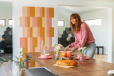 a felt right wall art design in shades of cream, yellow, and pink o a white wall in a dinning room with a woman setting the table