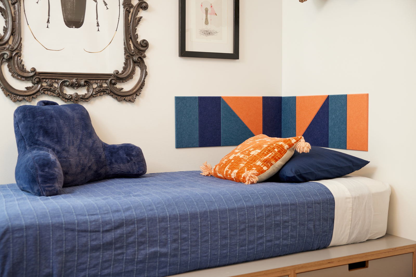 A Felt Right wall art design in shades of blue and orange in a corner of a bedroom above a bed.