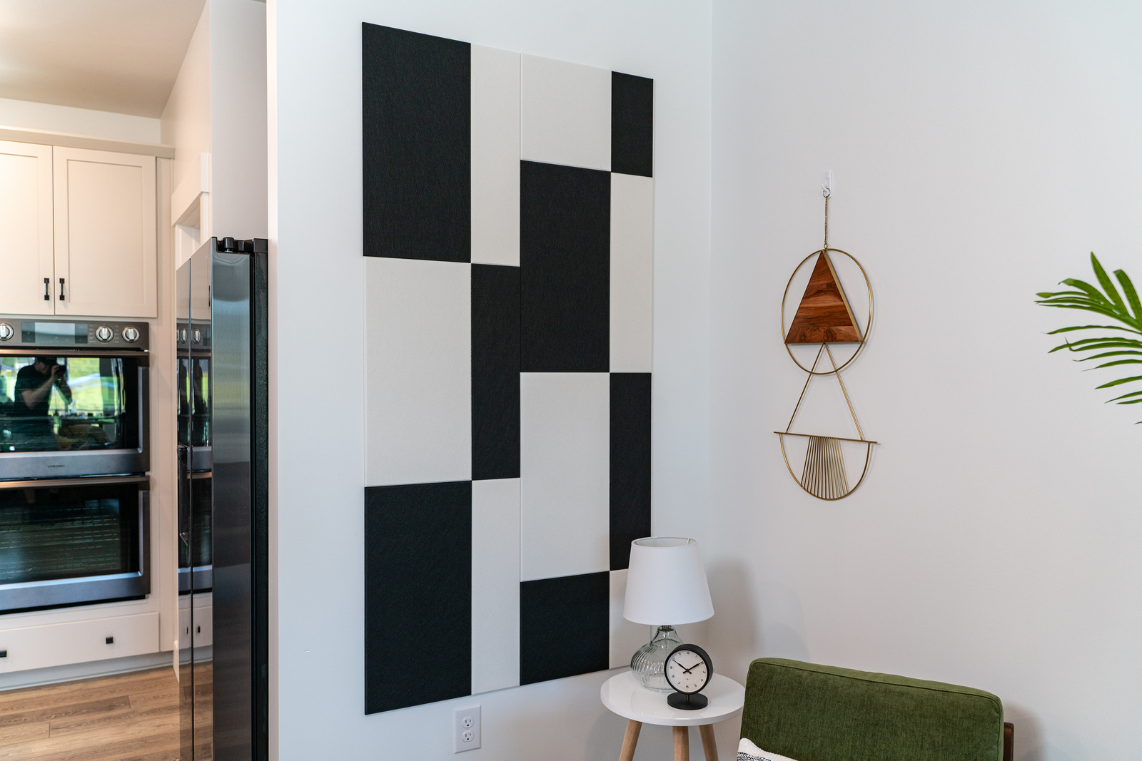 A Felt Right wall art design in black and white rectangles and squares on a white wall next to a kitchen and a side table with a little lamp.