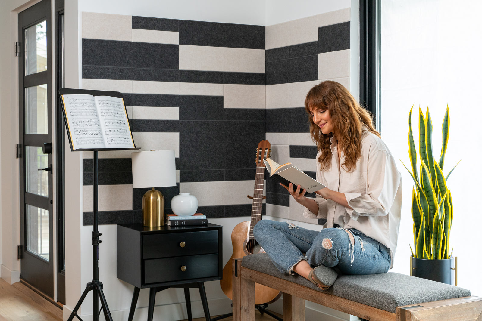 A Felt Right wall art design in shades of white and black felt tiles on a white wall in a music room with a girl reading on a bench.