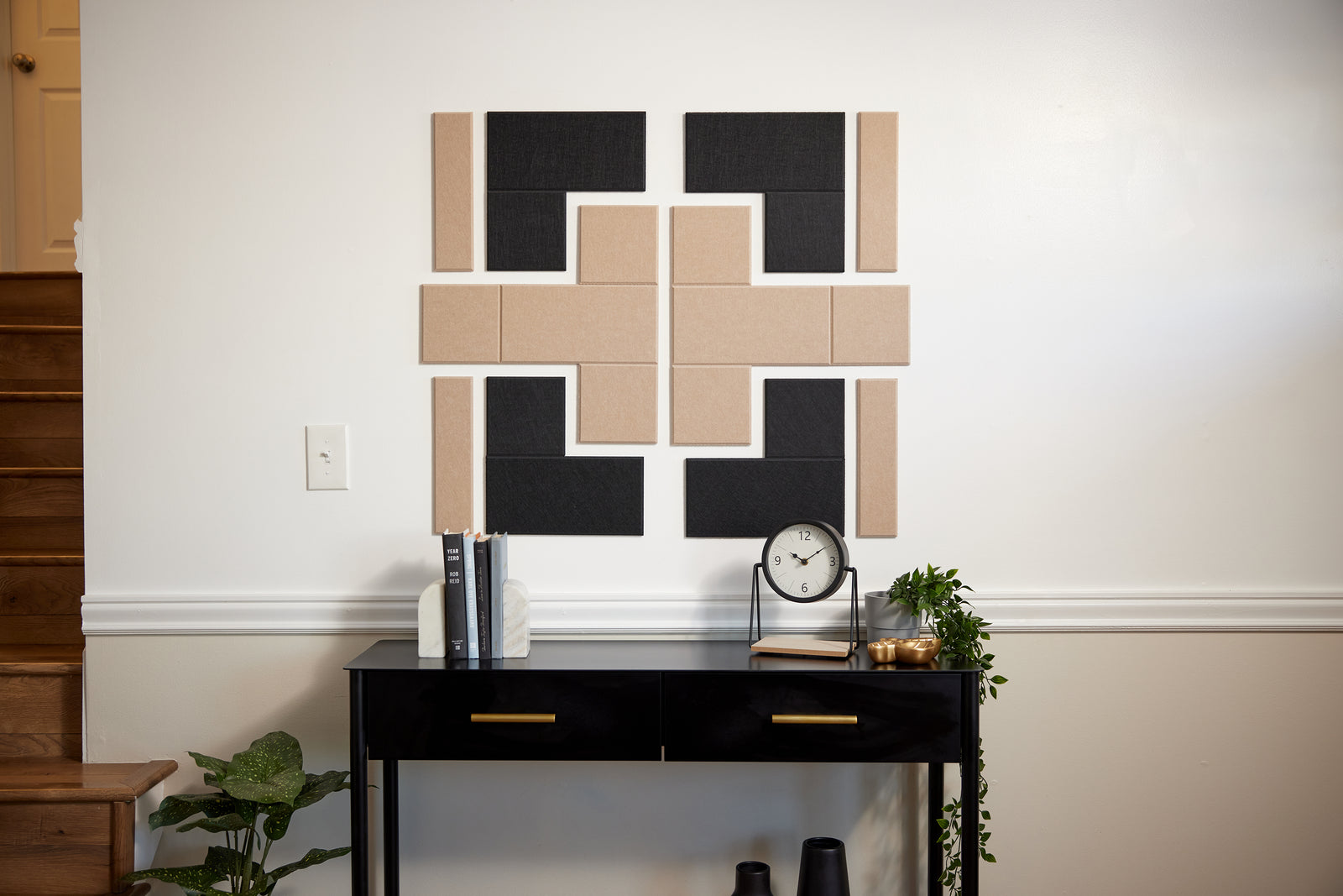 A Felt Right design on a wall above a table with books and a clock on it in shades of tan and black next to a stair case.