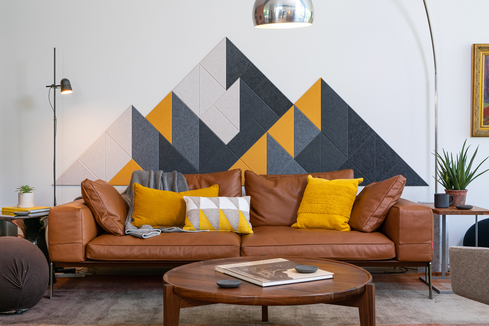 A Felt Right wall design wall decor featuring a geometric mountain-like pattern in shades of gray, black, and yellow, mounted on a wall above a brown sofa with yellow and gray throw pillows.