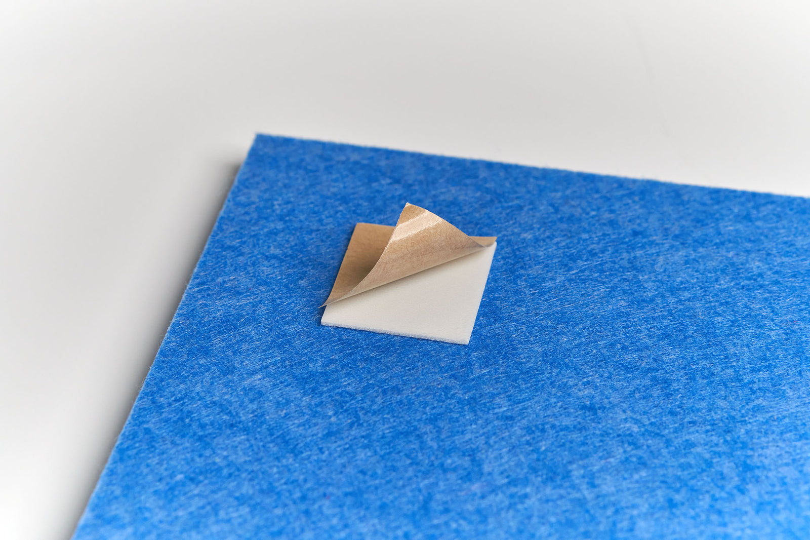 A Felt Right double-sided adhesive tab with its brown backing partially peeled back is placed on the back of a blue Felt Right tile.