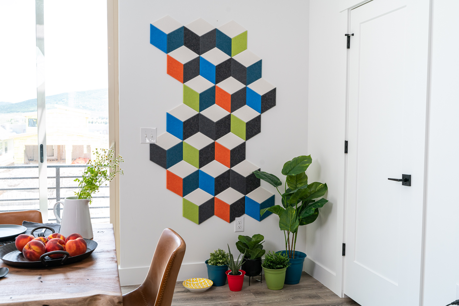 A Felt Right wall geometric art design in grey, blue, green, orange, and cream forming a 3d like cube design next to a door and some house plants.