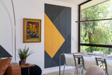 Felt Right wall art design in shades of yellow and black felt tiles on a white wall next to a flower painting and window located in a dinning room