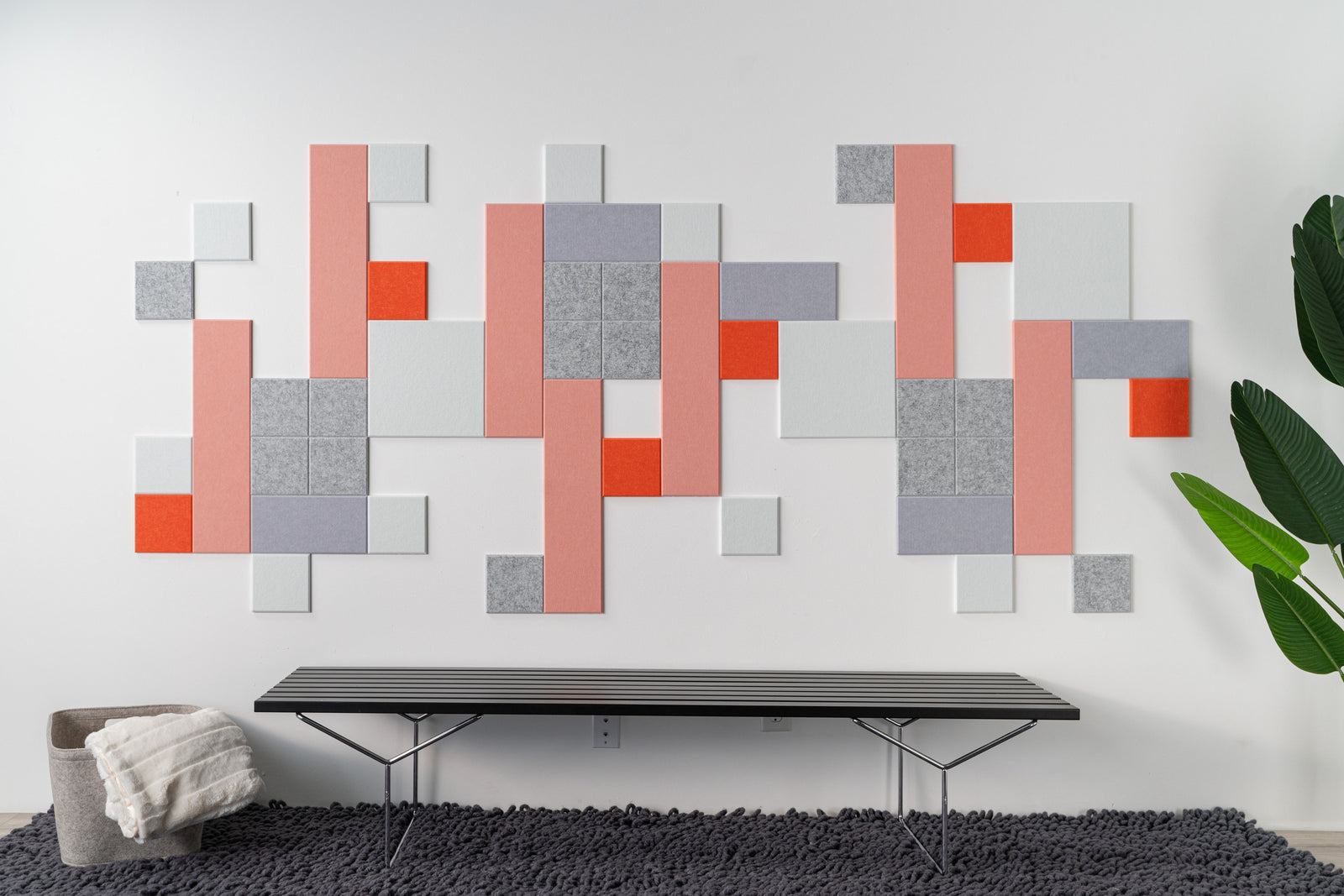 A Felt Right wall art design in shades of pink orange purple grey and white felt tiles on a white wall above a bench on a rug.
