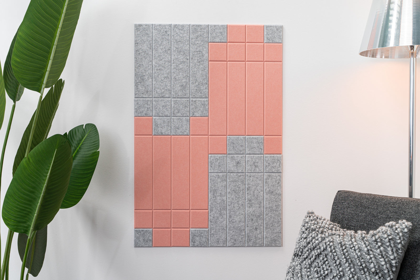 A Felt Right wall art design in shades of pink and grey felt tiles on a white wall next to a houseplant and a chair with a lamp behind it.