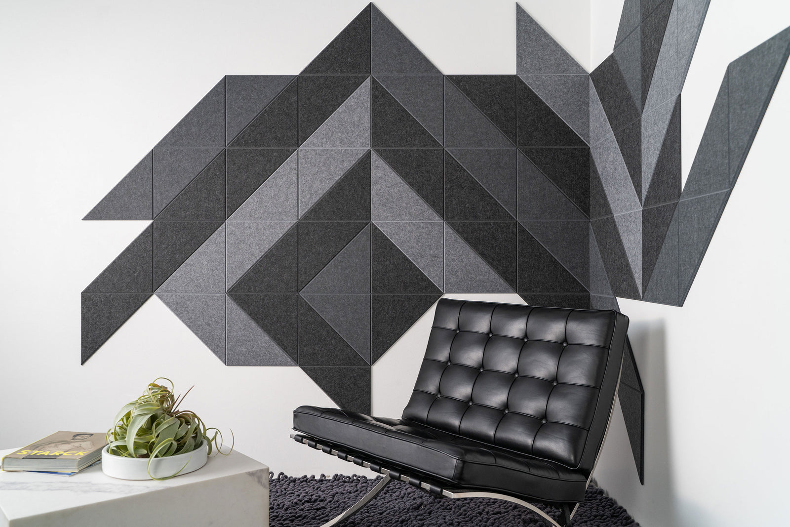 A Felt Right wall art design in shades of grey and black felt tiles on a white wall in a room with a black chair and a side table.