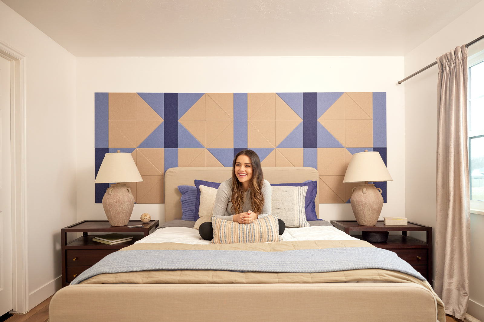 A Felt Right wall design in cream and blue behind a bed and two side tables with a woman sitting on the bed