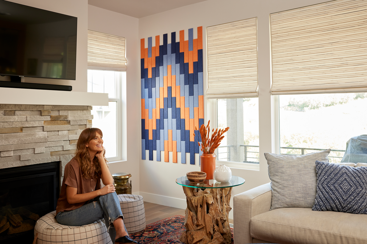 A Felt Right wall art design in shades of blue and orange in felt shiplap like tiles on a cream wall in between some windows in a living room with a fireplace.