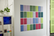 A Felt Right wall art design of rectangle tiles and negative space in shades of blues, greens, pinks, and purples on a wall with a shelf and plants next to it.