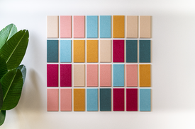 A Felt Right Mosaic wall design in shades of pink, blue, yellow, and cream displayed on a white wall with a plant in the corner