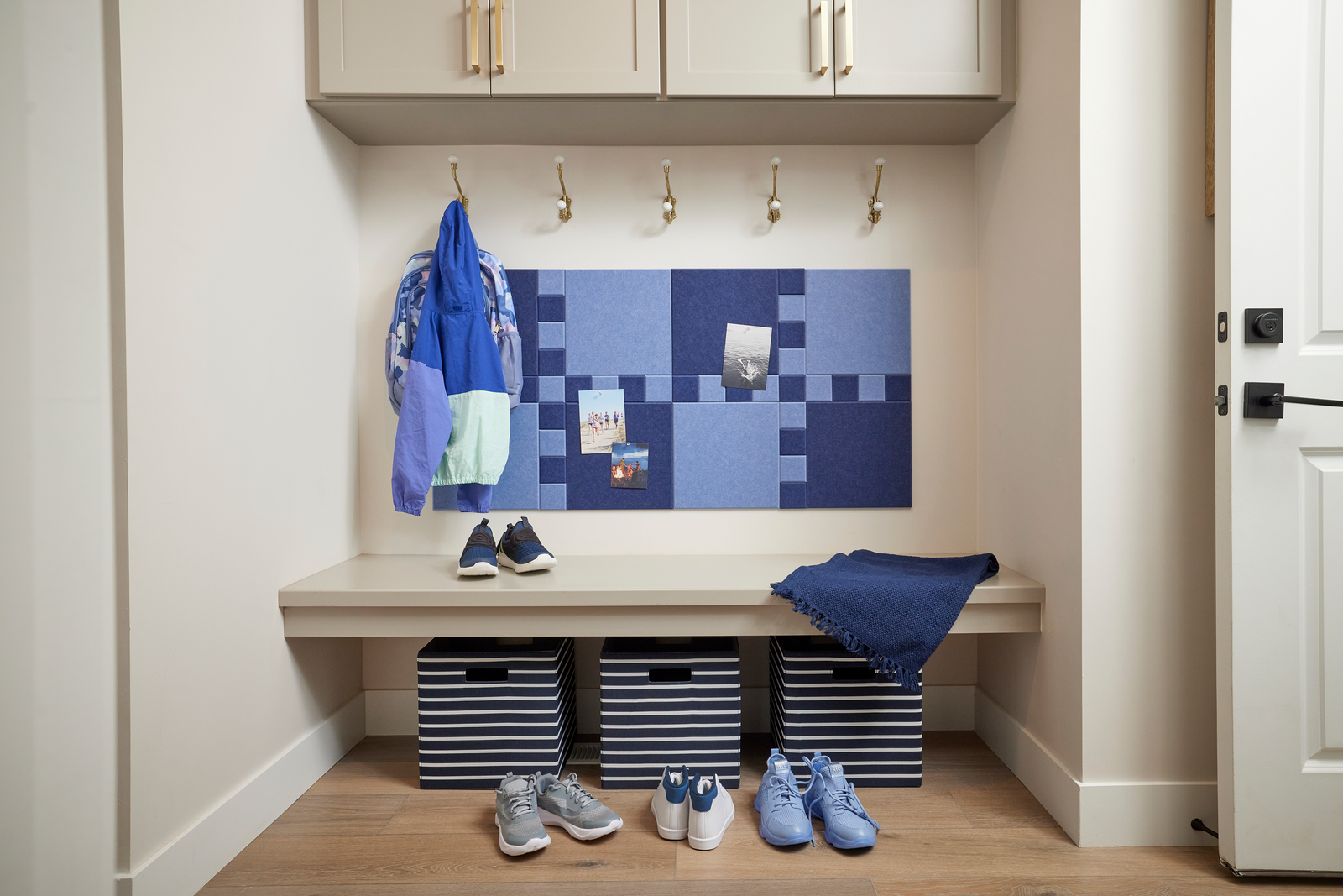 A Felt Right wall art design in shades of blue felt squares making a puzzle like pattern on a cream wall in a mudroom with coat hooks and a bench.