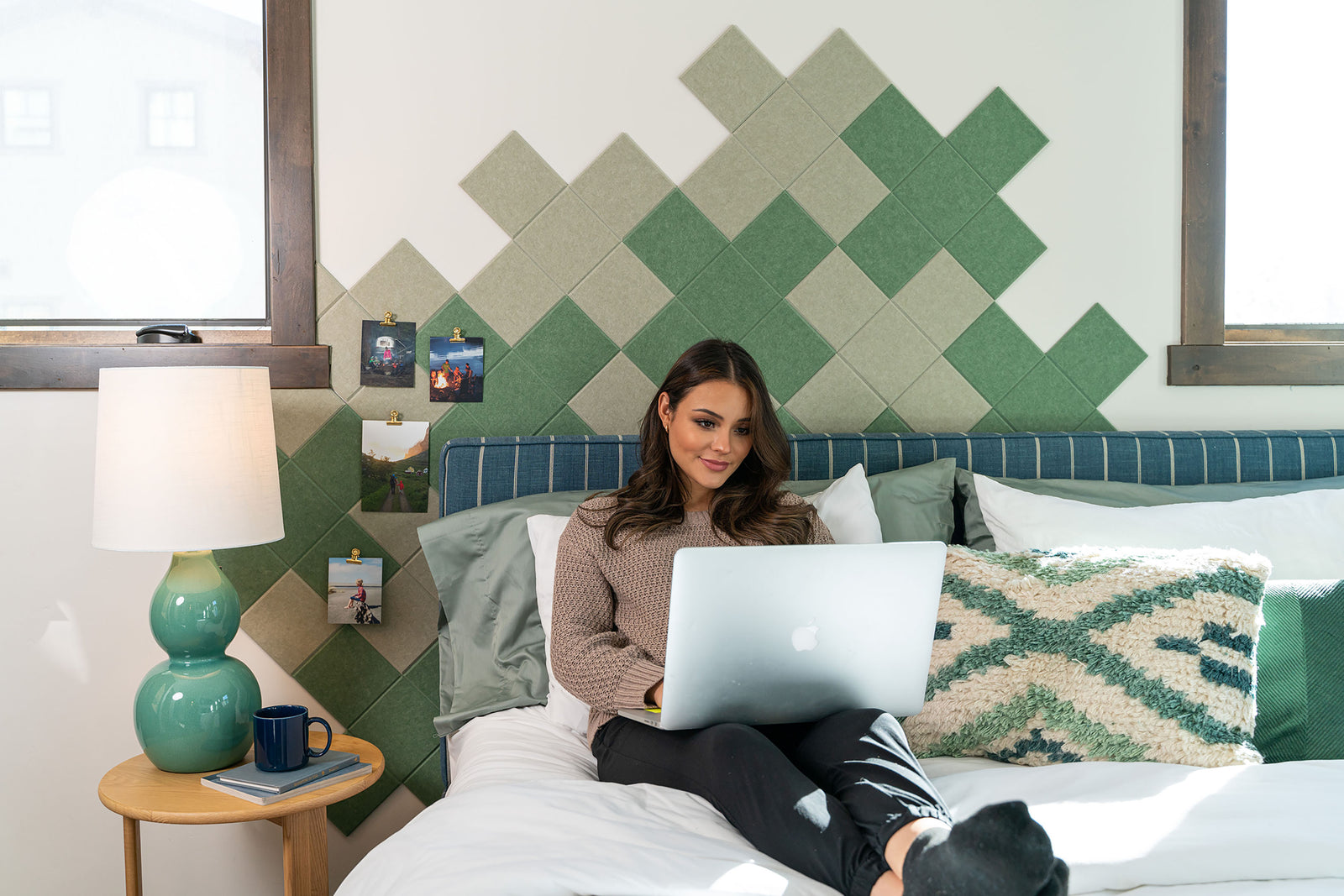 A Felt Right wall art design in shades of green felt tiles on a white wall with images pinned on it behind a bed with a woman working on her laptop.