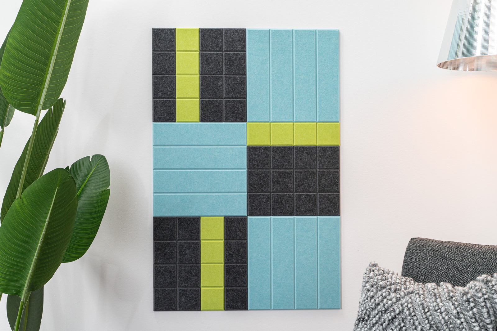 A Felt Right wall art design in shades of blue, green, and black felt tiles on a white wall with house plant  and a chair with a lamp behind it.