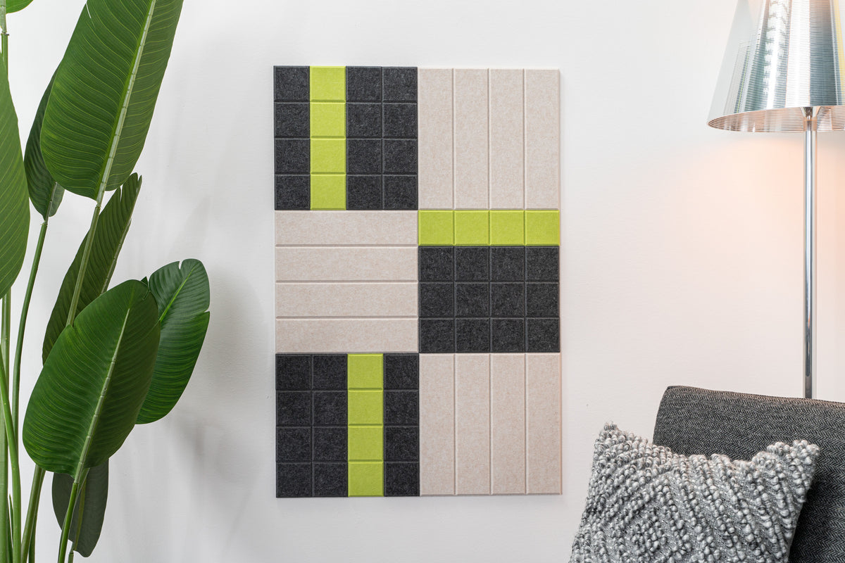 A Felt Right wall art design in shades of cream, green, and black felt tiles on a white wall with a houseplant and a chair with a lamp behind it.