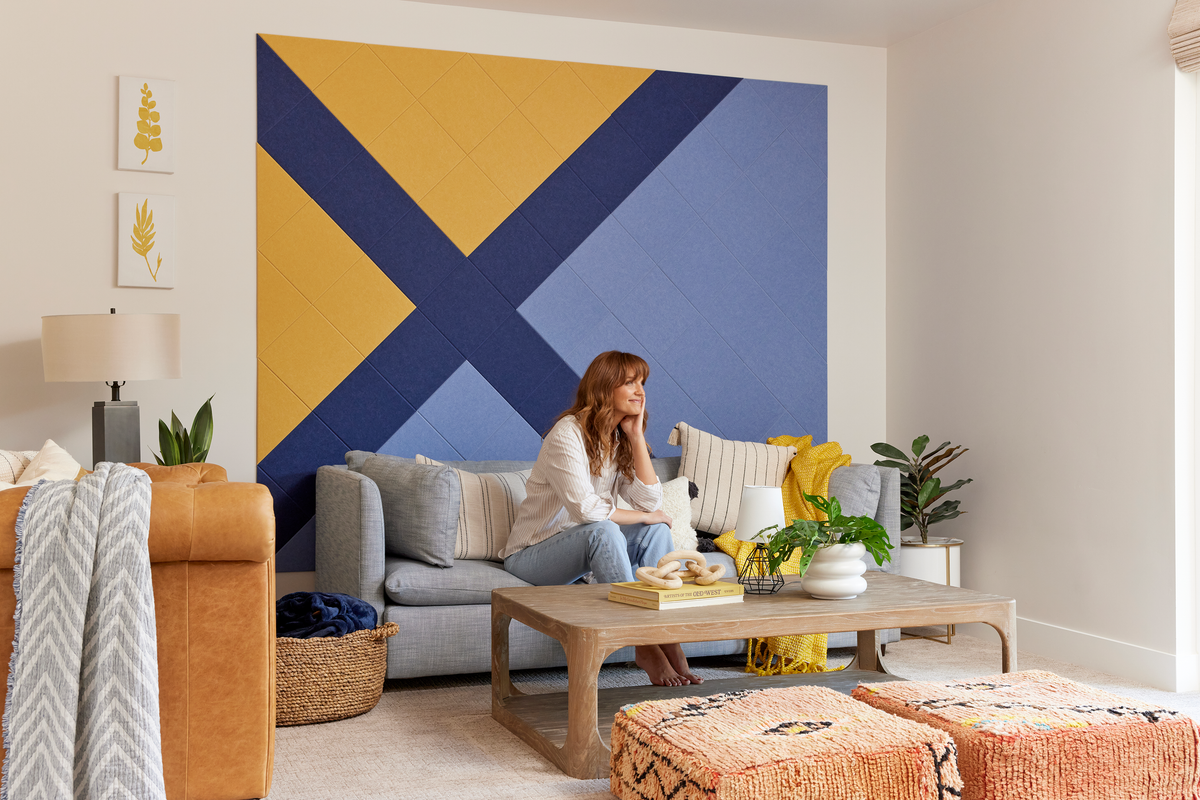 Felt Right wall art using shades of blue and yellow felt tiles on a white wall in a living room behind a couch with a woman sitting on it