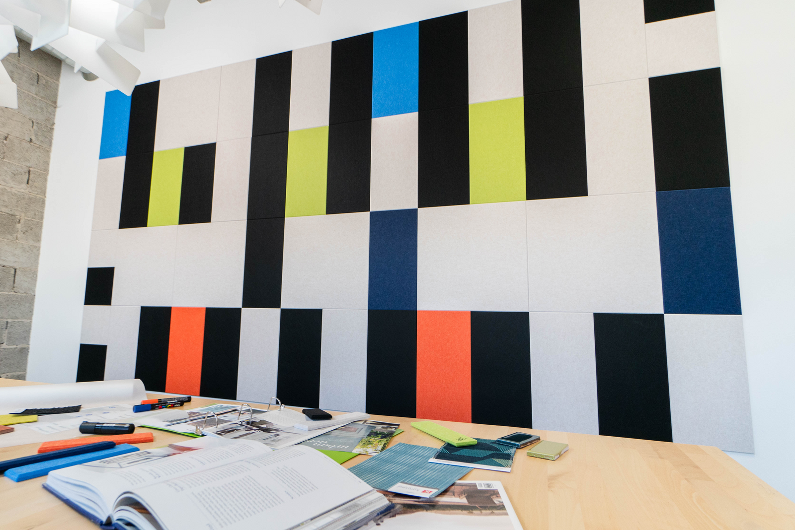 A Felt Right wall art design in shades of white, black, orange, blue, and green felt tiles on a white wall in a room with a big desk.