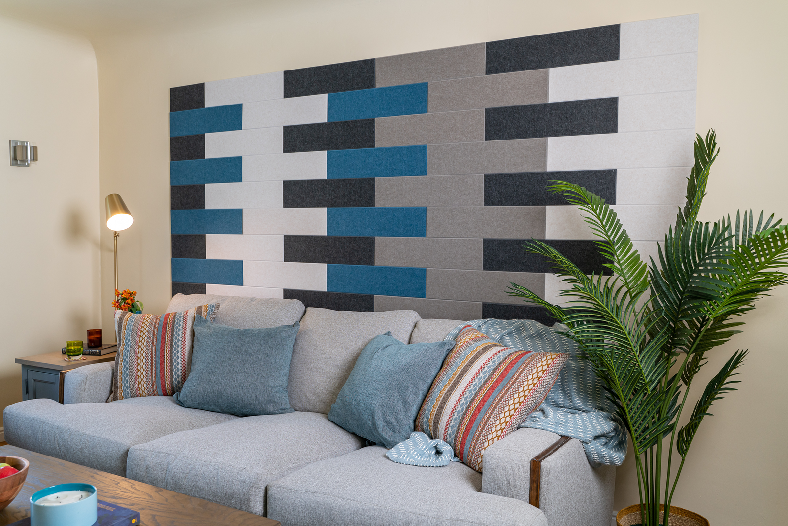 A Felt Right wall art design in shades of blue, white, grey, and black in felt tiles on a wall above a couch with decorative pillows.