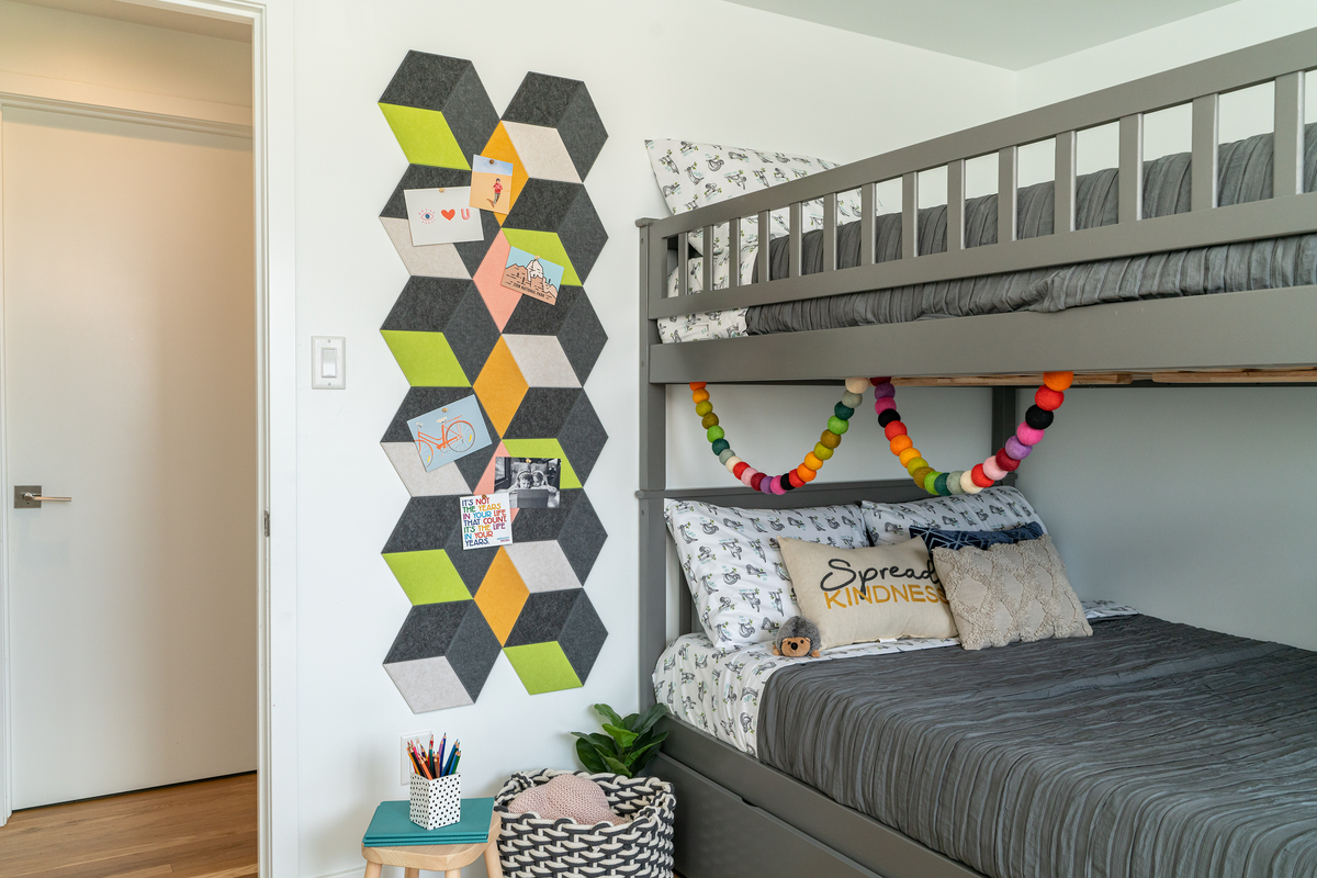 A Felt Right wall art design in shades of grey, green, yellow, pink, and white diamond shaped felt tiles on a white wall next to a bunkbed in a bedroom.