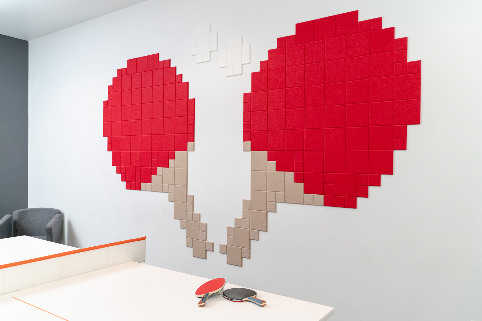 A Felt Right wall art design in shades of red brown and white felt tiles creating two ping pong paddles and two ping pong balls on a white wall with a ping pong table.