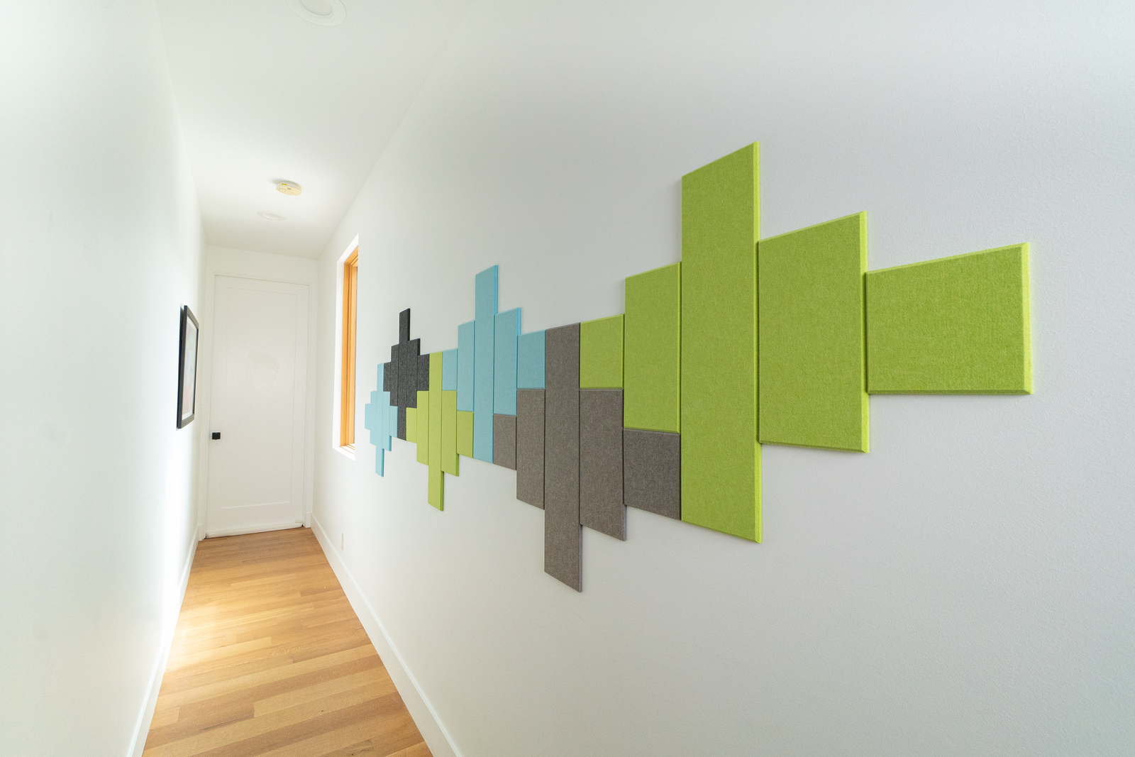 A Felt Right wall art design in blue, green, and grey in a geometric pattern along a white wall in a hallway.