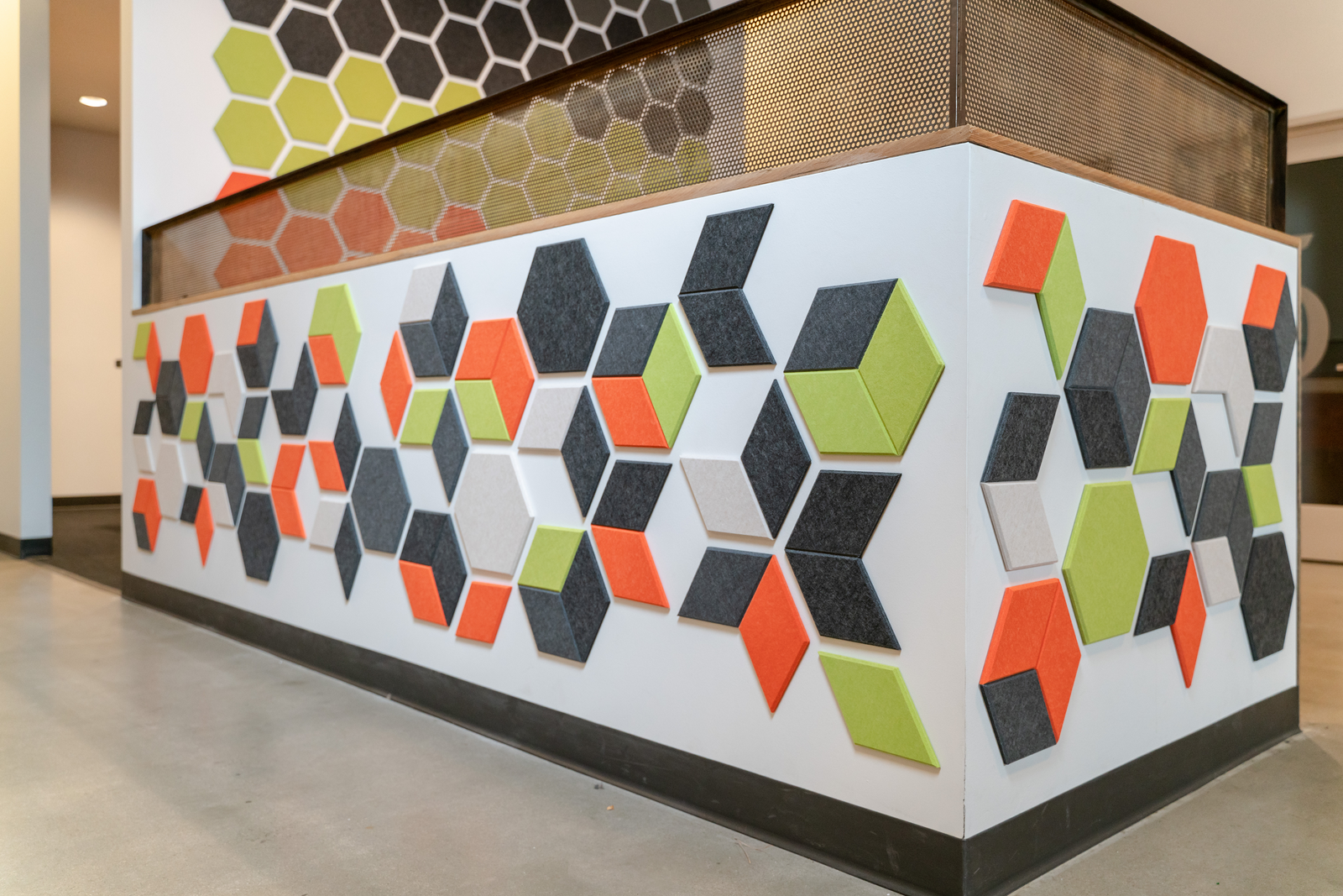 A Felt Right wall art design in shades of grey, green, orange, and white diamond felt tiles on a white wall that wraps around a corner in an office.
