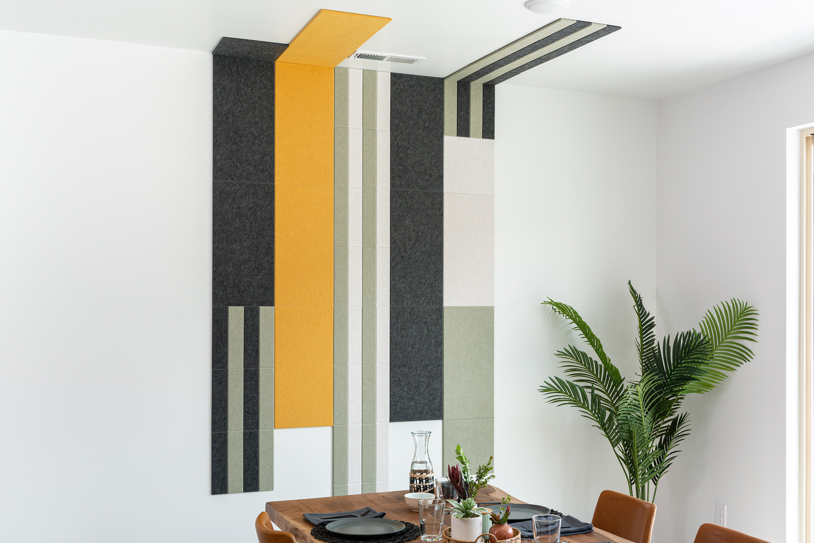 Felt Right wall art design in shades of green, yellow, white, and black on a white wall and ceiling in a dinning room with a house plant in the corner