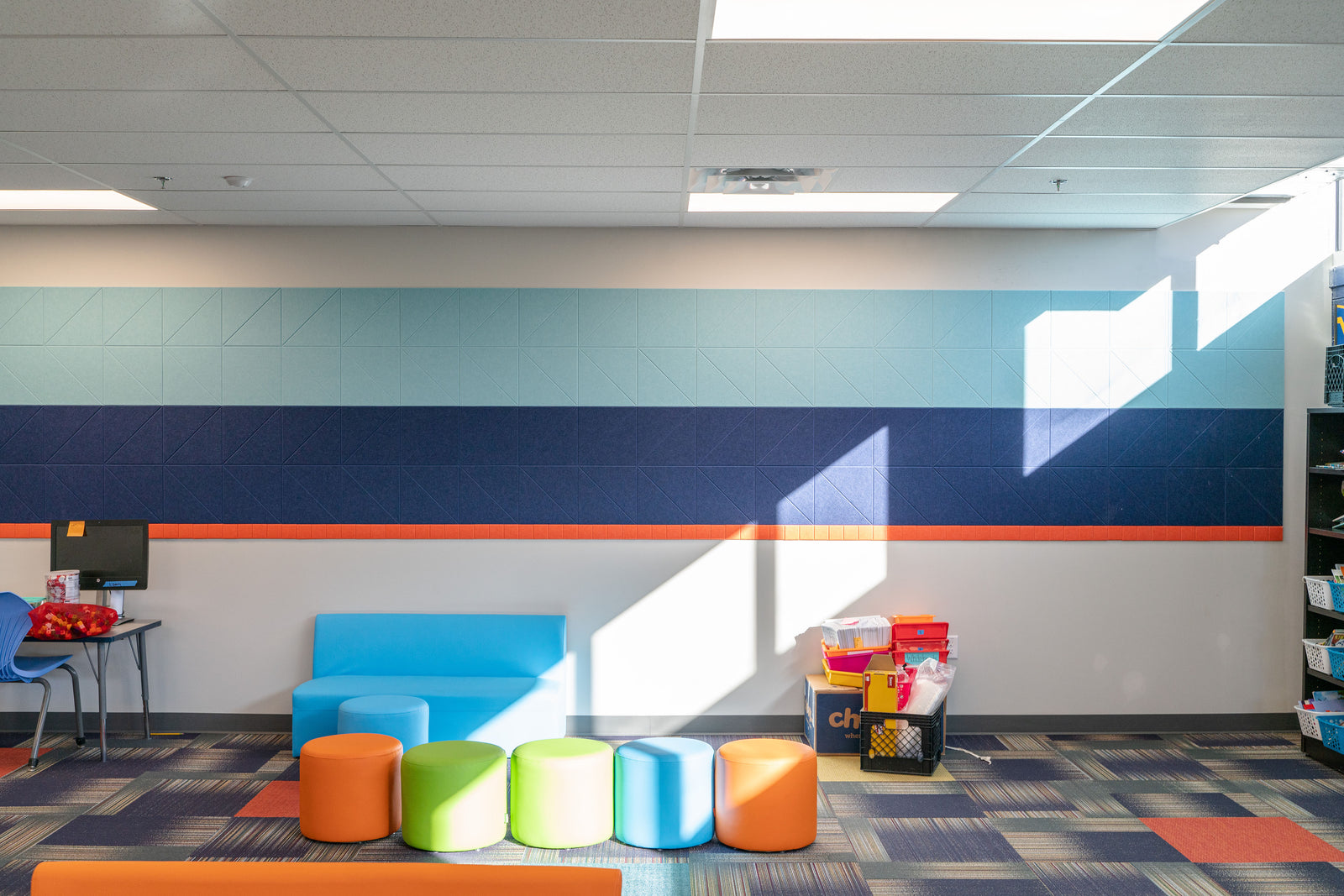 A Felt Right wall art design in shades of blue and orange felt tiles creating a strip along. a white wall in a classroom setting.