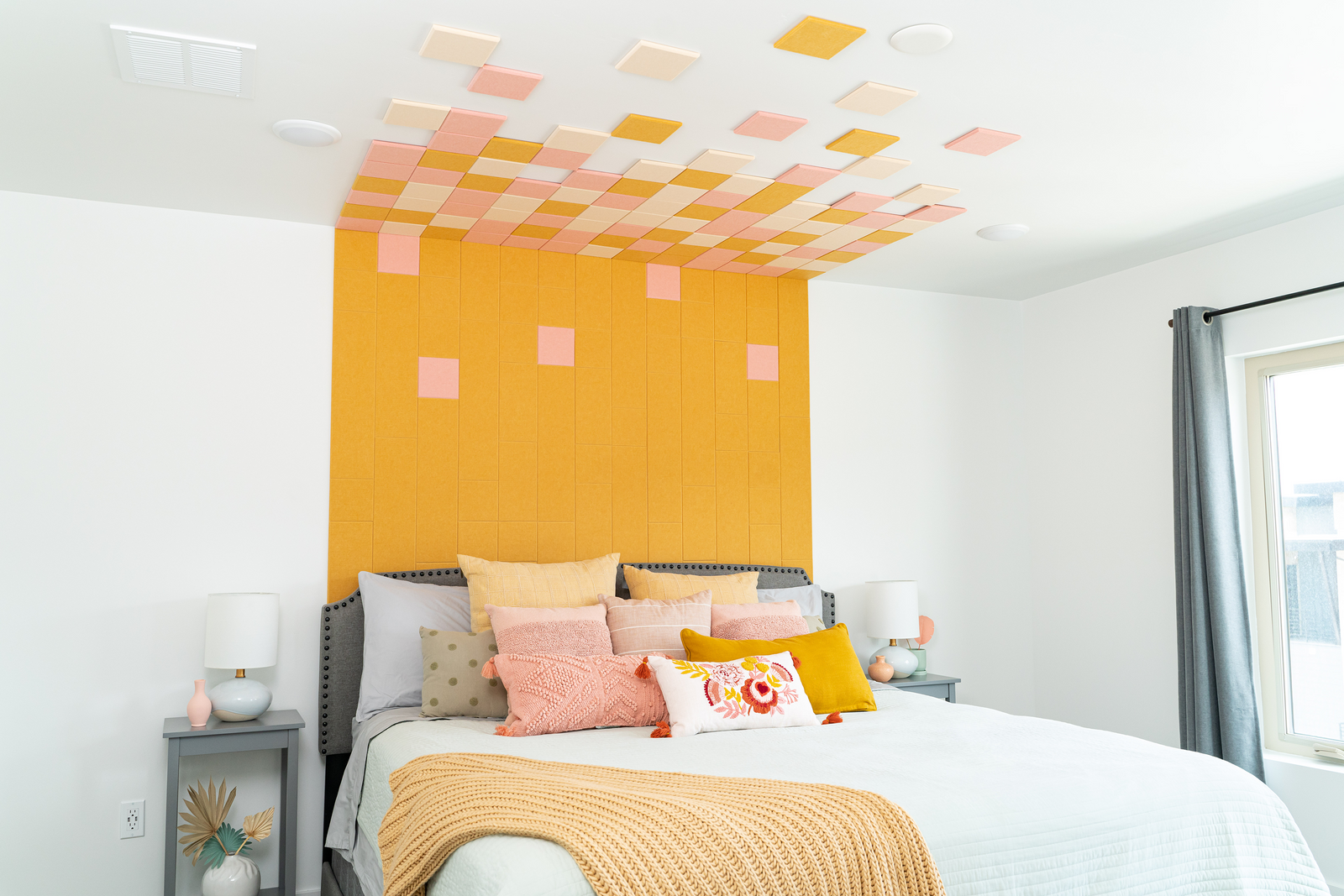 A Felt Right wall art design in shades of yellow, pink, and cream square felt tiles in a bedroom on a white wall and on the ceiling above a bed.