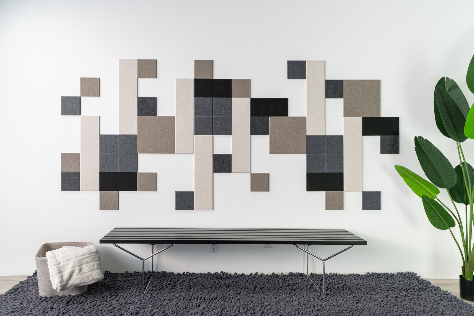 A Felt Right wall art design using shades of grey, cream, and black creating a geometric pattern above a bench on a rug next to a house plant.