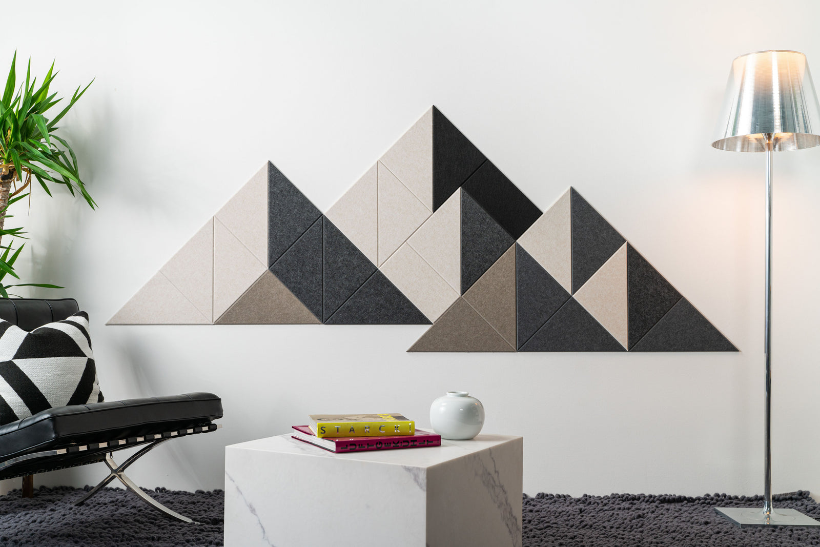 A Felt Right modern wall-mounted art piece resembling a mountain range, composed of triangular panels in shades of beige, gray, and black, installed above a white bench in a room with a floor lamp and a plant.